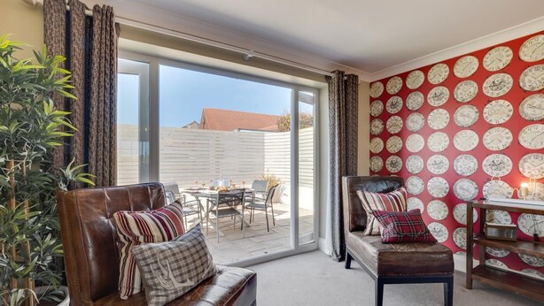 Een kamer met een leren bank, een eettafel met stoelen, een rode muur met klokken en een glazen schuifdeur naar een buitenruimte.