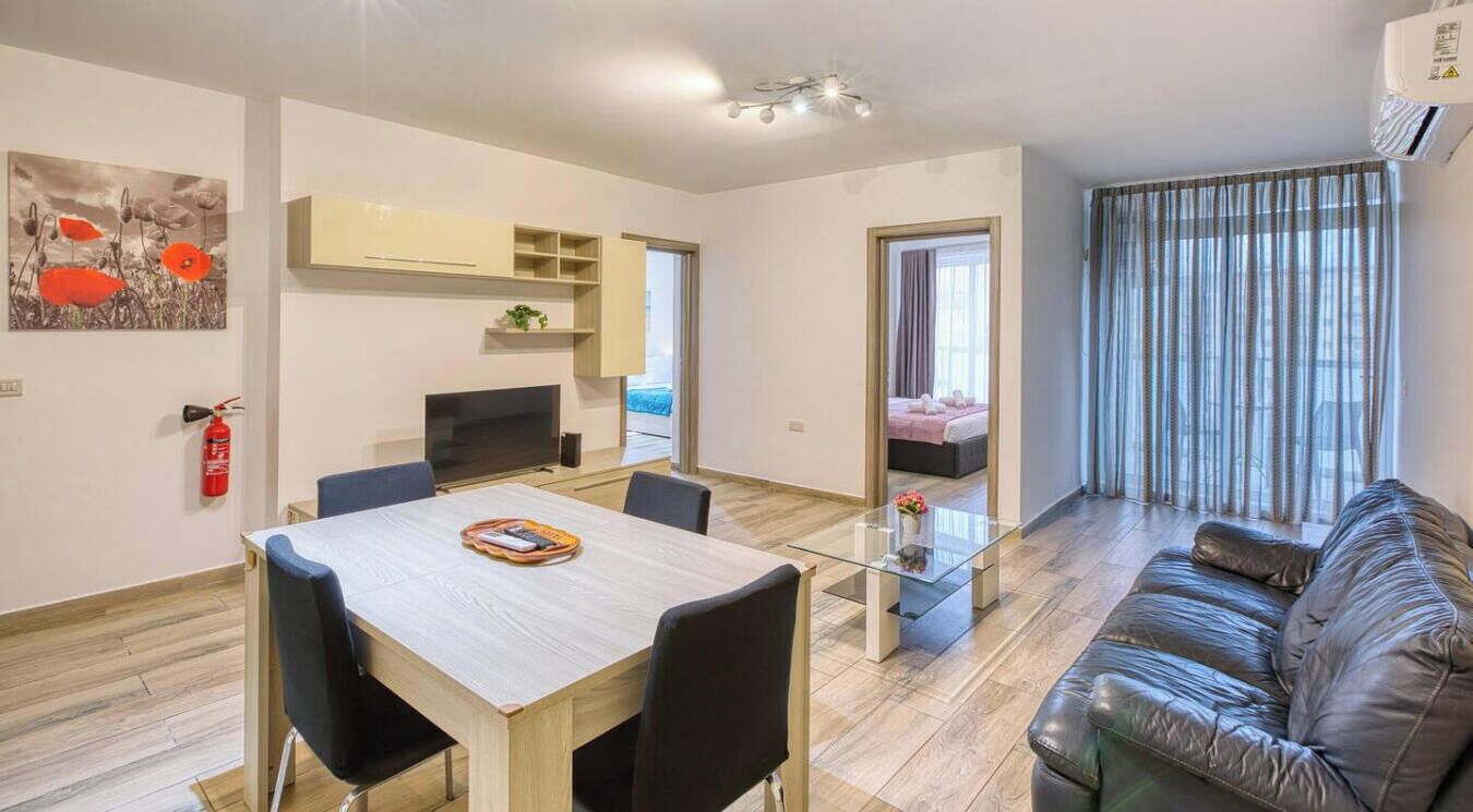 Een moderne woonkamer met een glazen salontafel, een leren bank, een eethoek met een houten tafel en stoelen, en een keuken met een televisie en kasten.