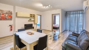 A modern living room with a glass coffee table, a leather sofa, a dining area with a wooden table and chairs, and a kitchen area with a TV and cabinets. - Soft Coastal Vibes with King Beds & Balcony! (St. Paul`s Bay)