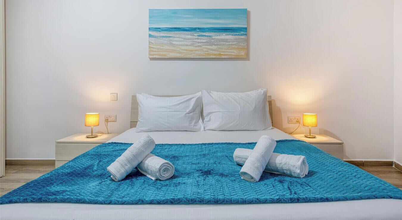 Een hotelkamer met een bed, twee bedlampen, een blauw tapijt met structuur en een schilderij van een strand.