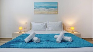 A hotel room with a bed, two bedside lamps, a blue textured rug, and a painting of a beach scene on the wall. - Soft Coastal Vibes with King Beds & Balcony! (St. Paul`s Bay)