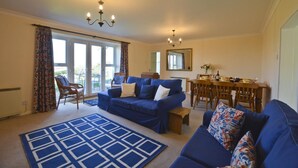 A living room with a blue sofa set, a dining table with chairs, a chandelier, and a large window with curtains. - 2 Bed in Morpeth (oc-nb596) (Low Hauxley)