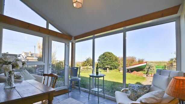 A sunlit room with a dining table, chairs, and a sofa. Large windows offer a view of a garden and a boat. - 2 Bed in Morpeth (oc-nb596) (Low Hauxley)