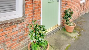 Een bakstenen huis met een groene deur waarop 'Mundles' staat, twee potplanten en een klein raam met een witte omlijsting.