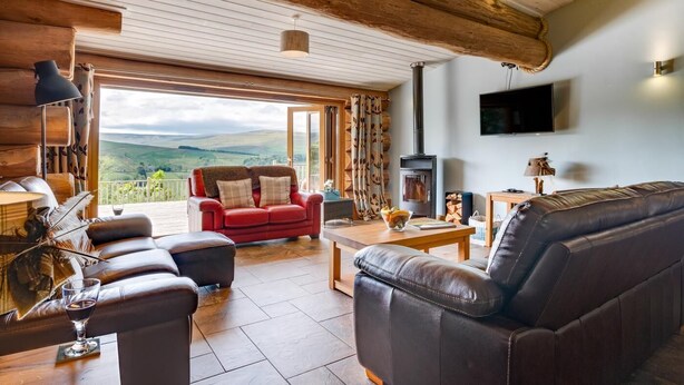 A cozy living room with a fireplace, leather sofas, and a large window offering a scenic view. - 4 Bed in Alston (oc-nb575) (Alston Moor)