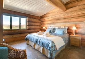 A log cabin bedroom with a large bed, two bedside tables with lamps, a wicker chair, and a view of the outdoors through a window.