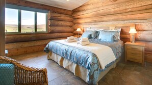 A log cabin bedroom with a large bed, two bedside tables with lamps, a wicker chair, and a view of the outdoors through a window. - 4 Bed in Alston (oc-nb575) (Alston Moor)