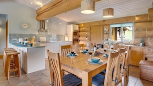 A modern kitchen with a central island, wooden dining table, and chairs. There is a clock on the wall and a window providing natural light. - 4 Bed in Alston (oc-nb575) (Alston Moor)