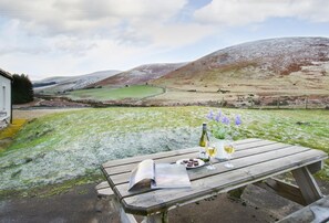 Une table de pique-nique en bois, avec une bouteille de vin, des verres et un menu, est placée sur une colline herbeuse, offrant une vue sur des collines ondulantes et un bâtiment en arrière-plan.