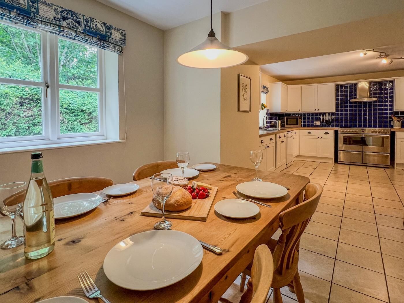 A dining area with a wooden table set for four, a bottle of wine, and a fruit platter. The room has a window with a view of greenery, a kitchen with blue tiles, and a hanging light fixture.