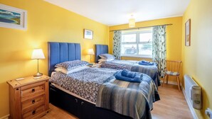 Ein Hotelzimmer mit zwei Betten, einem Holzkoffer, einem Nachttisch, einem Fenster mit Vorhängen und einer Heizung.