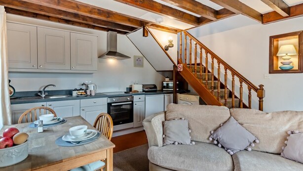 A cozy kitchen with wooden beams, a dining table with plates and apples, a sofa with pillows, and a staircase with a banister. - 1 Bed in Haltwhistle (oc-nb556) (Haltwhistle)