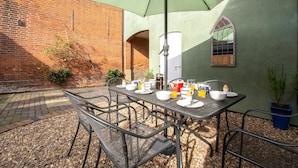 An outdoor patio with a table set for breakfast, surrounded by chairs, and an umbrella providing shade. - 3 Bed in Cromer (oc-1927) (Cromer)