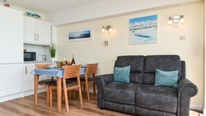 Een woonkamer met een donkere bank, houten stoelen, een eettafel met een blauwe tafelkleed en een keukenhoek met witte kastjes en een magnetron.