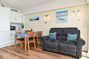 A living room with a dark sofa, wooden chairs, a dining table with a blue tablecloth, and a kitchen area with white cabinets and a microwave. - 2 Bed in Saundersfoot (oc-pw9131) (Saundersfoot)
