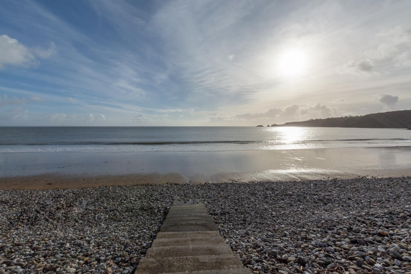 A pebbled beach with a concrete path leading to the water, under a partly cloudy sky.