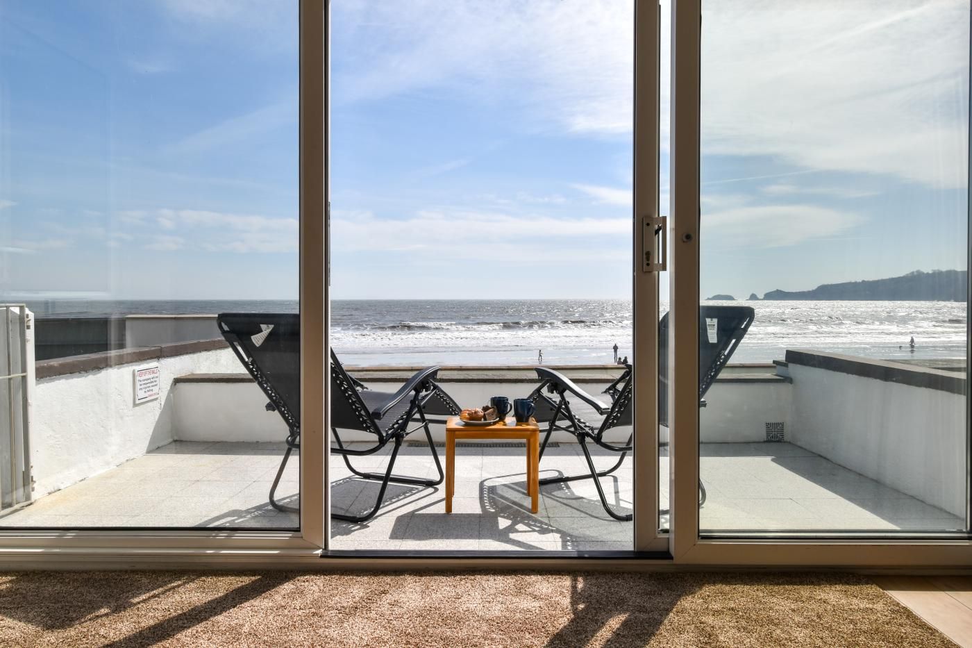 Un balcon avec deux chaises et une petite table, donnant sur une plage, l’océan et des collines lointaines.