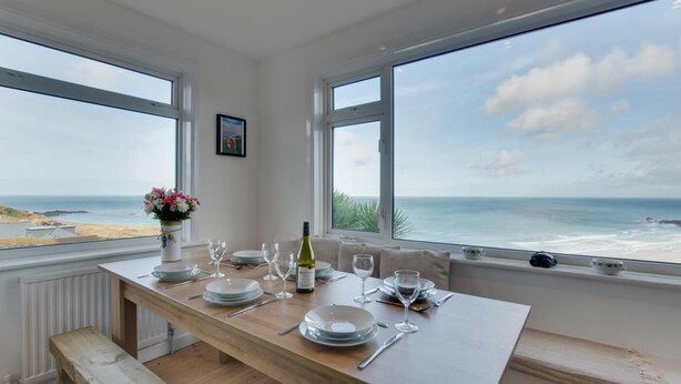 Un coin repas avec une table dressée pour quatre personnes, de grandes fenêtres donnant sur la mer et un tableau accroché au mur.