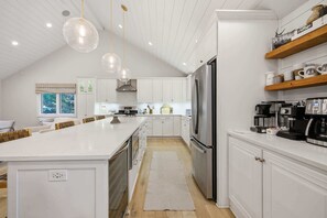 A modern kitchen with white cabinetry, a central island, stainless steel appliances, pendant lighting, and wooden shelves. - Loma House - Pool, Hot Tub & Sauna walk to Lake MI (Sawyer)
