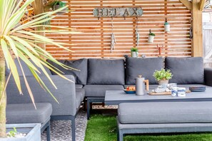 A modern outdoor seating area with a wooden slatted wall, a grey sofa, and a coffee table with various items. - 2 Bed in Padstow (oc-p00714) (Padstow)
