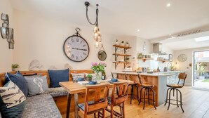 A cozy living room with a sofa, dining table, and chairs, a kitchen area with a bar, and a large wall clock. - 2 Bed in Padstow (oc-p00714) (Padstow)