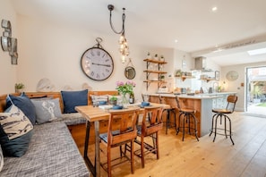 A cozy living room with a sofa, dining table, and chairs, a kitchen area with a bar, and a large wall clock. - 2 Bed in Padstow (oc-p00714) (Padstow)
