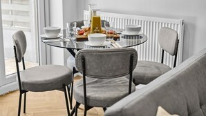 Un coin repas avec une table ronde en verre, des chaises à revêtement gris, un radiateur blanc et des aliments pour le petit-déjeuner.