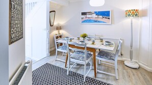 Un comedor con una mesa dispuesta para cuatro personas, sillas blancas con cojines estampados, un cuadro sobre la mesa y una lámpara de pie a la derecha.