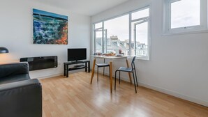 Ett modernt vardagsrum med en platt-TV, en tavla, en matplats med bord och stolar samt stora fönster med utsikt över staden.