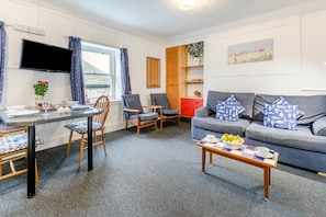A cozy living room with a grey sofa, a dining table with chairs, a TV mounted above the table, and a window with curtains. - 1 Bed in St Ives (oc-cher4) (Saint Ives)
