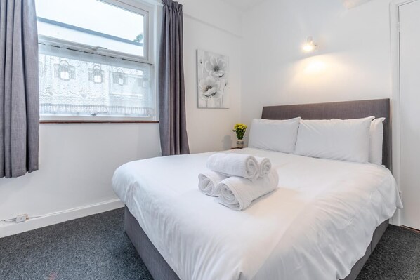 A neatly made bed with white linens and rolled towels, a window with curtains, a wall-mounted light, and a small vase with flowers. - 1 Bed in St Ives (oc-cher4) (Saint Ives)