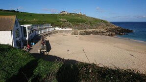 Un edificio costero con una playa en primer plano, una colina cubierta de hierba con una estructura en la cima y el mar al fondo.
