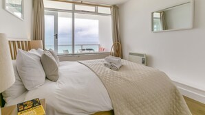 Een hotelkamer met een bed, uitzicht op zee, een raam met gordijnen, een nachtkastje met een boek en een spiegel aan de muur.