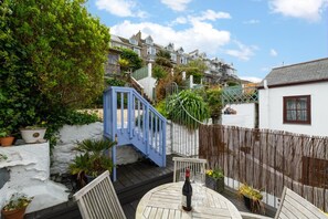 A patio with a table, chairs, a bottle of wine, and a blue staircase leading to a white house. - 3 Bed in St Ives (oc-twp21) (St Ives)