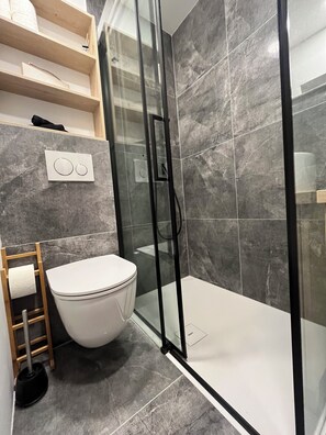 Un baño moderno con ducha de vidrio, inodoro y estante de madera con artículos de aseo.