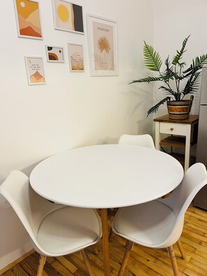 Ein runder, weißer Tisch mit vier weißen Stühlen, eine Zimmerpflanze auf einem hölzernen Beistelltisch und mehrere gerahmte Kunstwerke an der Wand.
