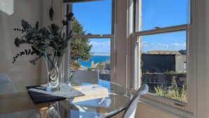 Spiseplads med et glasbord, hvide stole og en vase med grønne planter. Store vinduer giver udsigt til havet og bygninger.