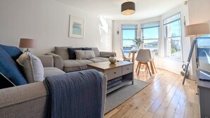 En moderne stue med sofa, sofabord og spiseplads med stole. Store vinduer giver udsigt udad.