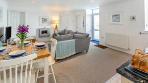 A modern living room with a fireplace, a dining area with a table set for two, and a kitchen with a countertop. - 2 Bed in St Ives (oc-trev2) (Saint Ives)