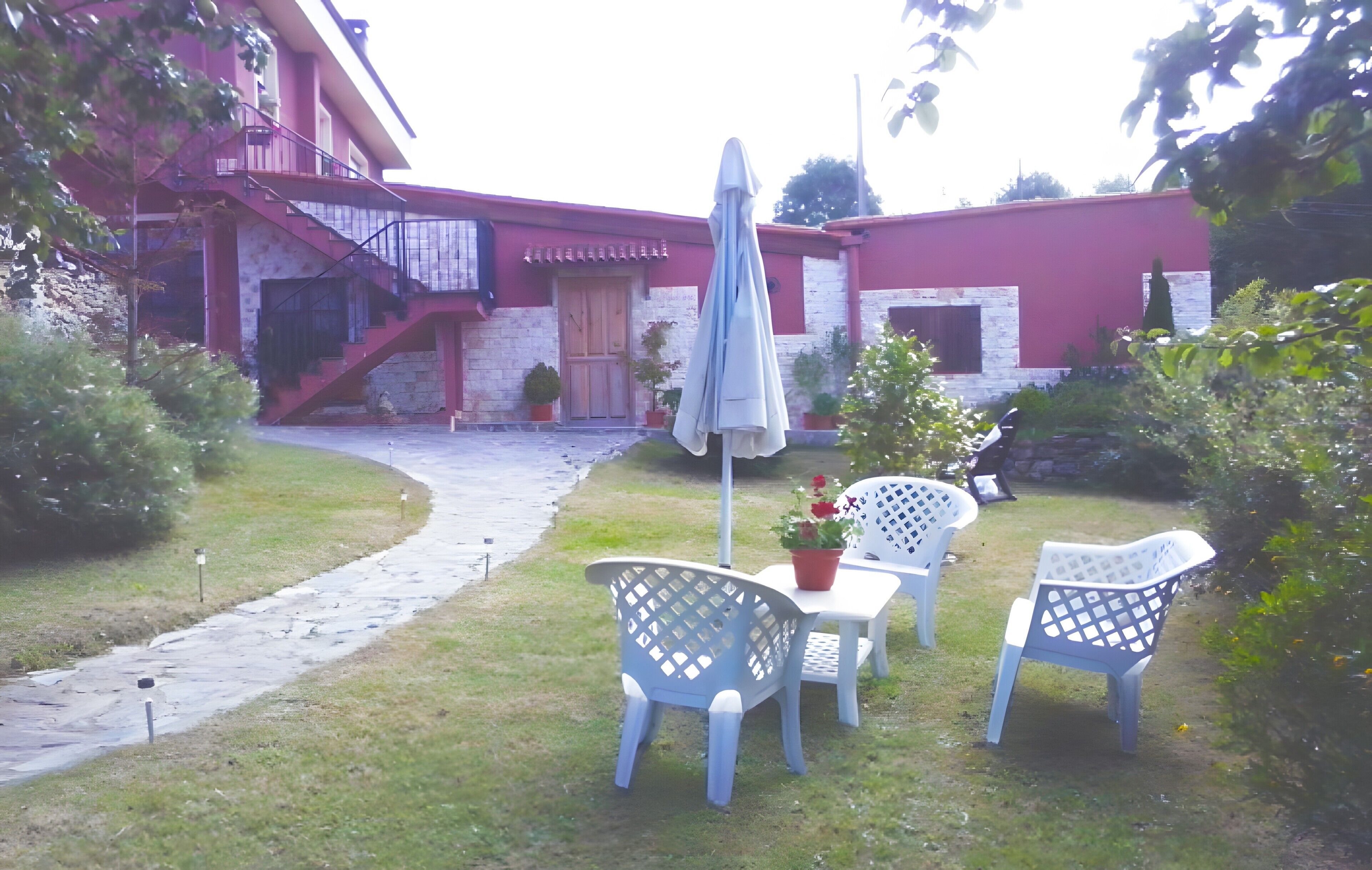Un edificio de fachada roja, una escalera de piedra y un camino pavimentado que conduce a un patio con mobiliario exterior y plantas en macetas.