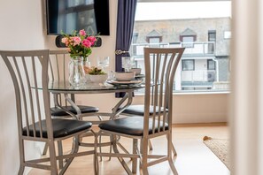Un coin repas avec une table en verre, des chaises en métal, un vase de fleurs et une vue sur les bâtiments par la fenêtre.