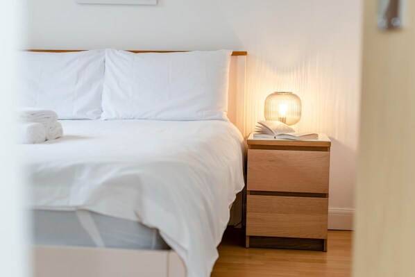 Un lit bien fait, avec du linge de lit blanc et une serviette pliée, une table de chevet avec une lampe et une petite table en bois à côté du lit.