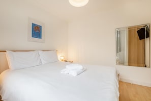 A neatly made bed with white linens and a folded towel, a bedside lamp, a framed picture on the wall, and a large mirror reflecting part of the room. - 1 Bed in St Ives (oc-clear2) (Saint Ives)