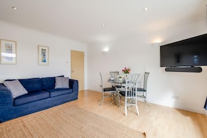A living room with a blue sofa, a dining table with chairs, a flat-screen TV mounted on the wall, and a wooden floor. - 1 Bed in St Ives (oc-clear2) (Saint Ives)