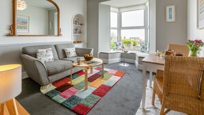 A living room with a grey sofa, a coffee table, a colorful rug, a wicker chair, and a window with a view. - 1 Bed in St Ives (oc-barn27) (Saint Ives)
