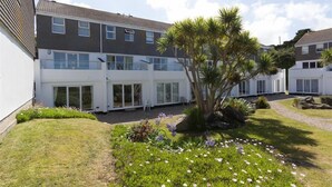 Un immeuble résidentiel moderne à deux étages, doté d’un patio couvert, d’un petit jardin agrémenté de fleurs et d’un palmier, et surplombant un ciel dégagé.