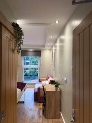 A narrow hallway with a wooden bench, a small table, and a window with a view of greenery. - Kastay: Bright London Studio | 1 Min to Trains (London)
