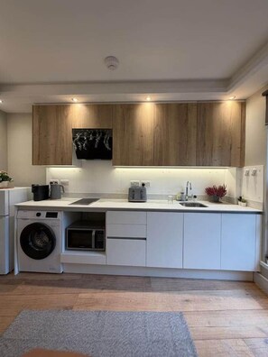 A modern kitchen with wooden cabinets, a built-in washing machine, and a grey rug on the floor. - Kastay: Bright London Studio | 1 Min to Trains (London)