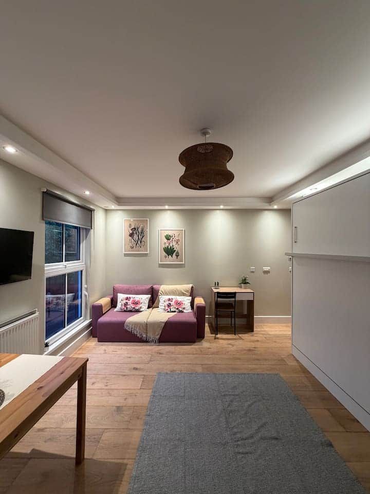 A modern living room with a sofa, a small table, a TV mounted on the wall, and a window.