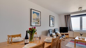 Un coin repas avec une table en bois dressée pour quatre personnes, une bouteille de vin et un vase de fleurs. Deux tableaux encadrés ornent le mur, ainsi qu’une télévision et une grande fenêtre avec des rideaux.
